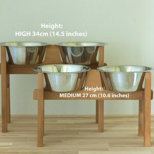 May include: A wooden pet feeder with two tiers, each holding two stainless steel bowls. The top tier is 34cm (14.5 inches) high, and the bottom tier is 27 cm (10.6 inches) high. The text on the image reads "Height: HIGH 34cm (14.5 inches)" and "Height: MEDIUM 27 cm (10.6 inches)".