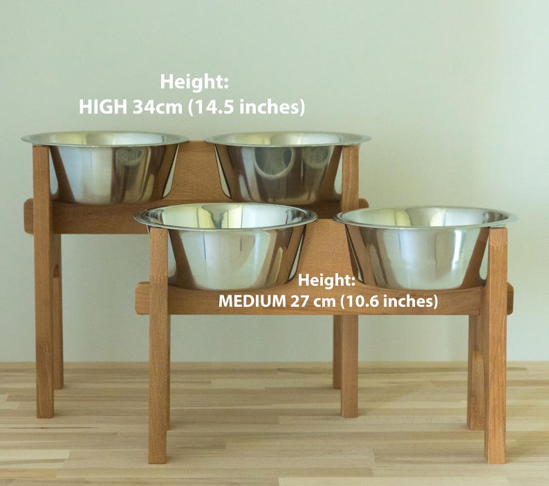 May include: A wooden pet feeder with two tiers, each holding two stainless steel bowls. The top tier is 34cm (14.5 inches) high, and the bottom tier is 27 cm (10.6 inches) high. The text on the image reads "Height: HIGH 34cm (14.5 inches)" and "Height: MEDIUM 27 cm (10.6 inches)".