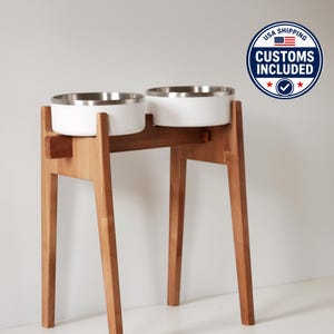 Peut inclure: Un support surélevé pour animaux de compagnie avec deux bols blancs et des rebords en acier inoxydable. Les bols reposent sur un support en bois naturel avec des pieds inclinés. Le support est conçu pour surélever les bols. Badge "USA Shipping".
