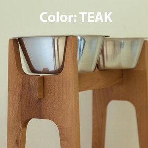 May include: Teak-colored wooden pet feeder with two stainless steel bowls. The feeder has a modern design with a sturdy base and a curved support for the bowls. The text "Color: TEAK" is visible in the image.