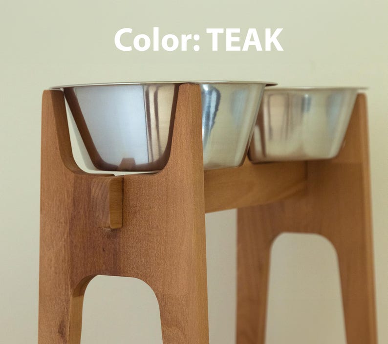 May include: Teak-colored wooden pet feeder with two stainless steel bowls. The feeder has a modern design with a sturdy base and a curved support for the bowls. The text "Color: TEAK" is visible in the image.