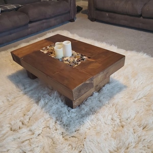 Op de afbeelding: Een rechthoekige salontafel van donker gebeitst hout. Het midden van de tafel is gevuld met kleine stenen en drie witte kaarsen. De tafel staat op een zacht, wit tapijt. Afmetingen: ongeveer 100 cm x 60 cm.