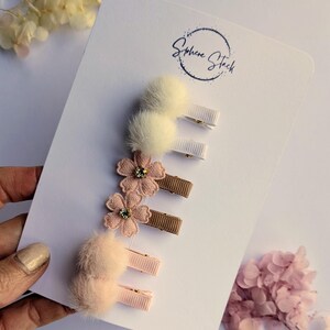 May include: A selection of hair clips presented on a white card. The clips showcase fluffy pom-poms, floral embellishments with rhinestones, and ribbon bows. The colour scheme includes cream, pink, and brown.