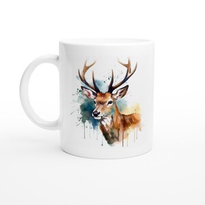 Könnte beinhalten: Weiße Keramiktasse mit einer Aquarell-Illustration eines Hirsches mit Geweih. Der Hirsch ist braun mit einem weißen Gesicht und ist von blauen und gelben Aquarellspritzern umgeben.