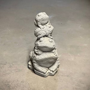 May include: A stack of three gray concrete frog figurines. The frogs are stacked on top of each other, with the largest frog at the bottom and the smallest frog at the top. The frogs are all facing forward, with their eyes closed.