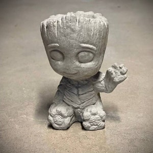 May include: A gray concrete planter shaped like a small, friendly character with a wide smile and large eyes. The planter has a pot on top of its head and one arm raised in a wave.