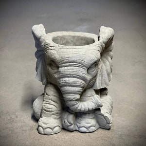 Peut inclure: Un pot en béton gris en forme d'éléphant avec une surface texturée. Le pot a une ouverture ronde en haut pour la plantation.