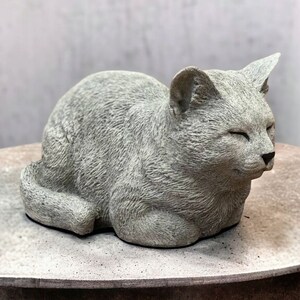 May include: A gray concrete sculpture of a cat sleeping with its eyes closed. The sculpture is detailed with fur and facial features.