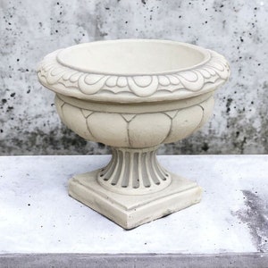 May include: A white ceramic planter with an ornate design around the top rim. The planter has a wide, round base and a tall, slender body.