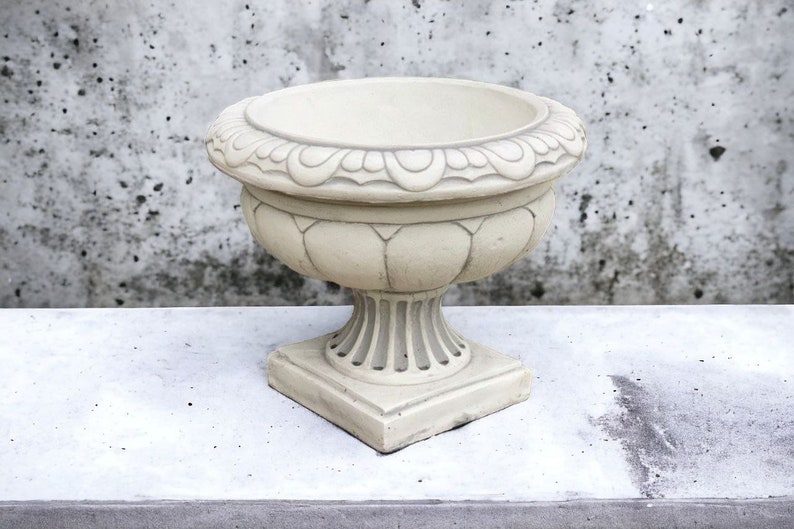 May include: A white ceramic planter with an ornate design around the top rim. The planter has a wide, round base and a tall, slender body.
