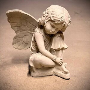 May include: A small, white, stone sculpture of a fairy with wings. The fairy is sitting with her head resting on her hand, looking down.