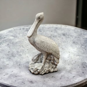 May include: A gray concrete pelican statue with a long beak and detailed feathers. The statue is perched on a rock base and is a decorative garden ornament.