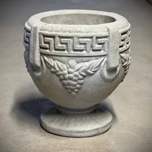 May include: A gray concrete planter with a Greek key design and grapevine accents. The planter has a round body and a pedestal base.