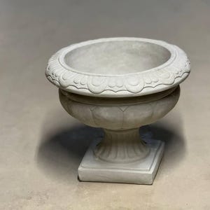 May include: A white, decorative, round planter with a fluted base and a detailed rim. The planter is made of concrete and has a classic design.