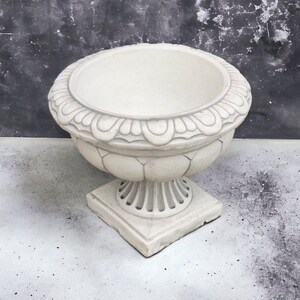 May include: A white ceramic planter with an ornate design around the top rim and a square base. The planter is empty and sits on a light gray surface.
