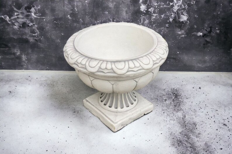 May include: A white ceramic planter with an ornate design around the top rim and a square base. The planter is empty and sits on a light gray surface.