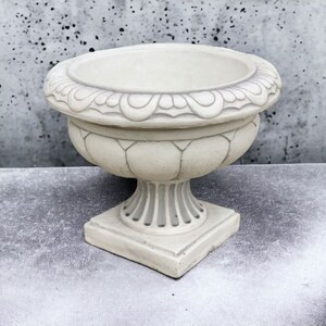 May include: A white ceramic planter with an ornate design around the top rim and a fluted design on the body. The planter has a square base and a round top.
