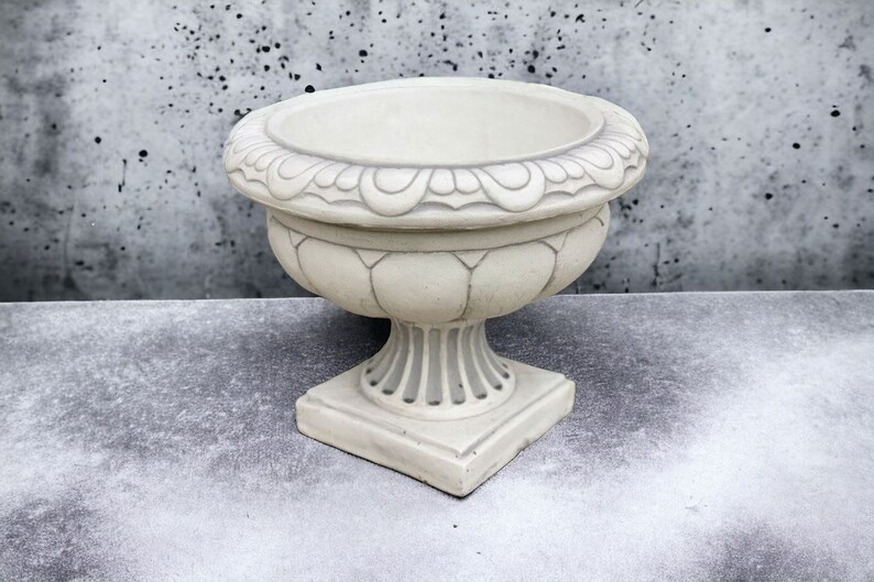 May include: A white ceramic planter with an ornate design around the top rim and a fluted design on the body. The planter has a square base and a round top.