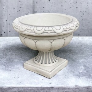 May include: A white ceramic planter with a decorative rim and a pedestal base. The planter is empty and ready for planting.