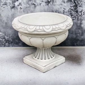 May include: A white ceramic planter with a decorative rim and a square base. The planter is sitting on a gray concrete surface.