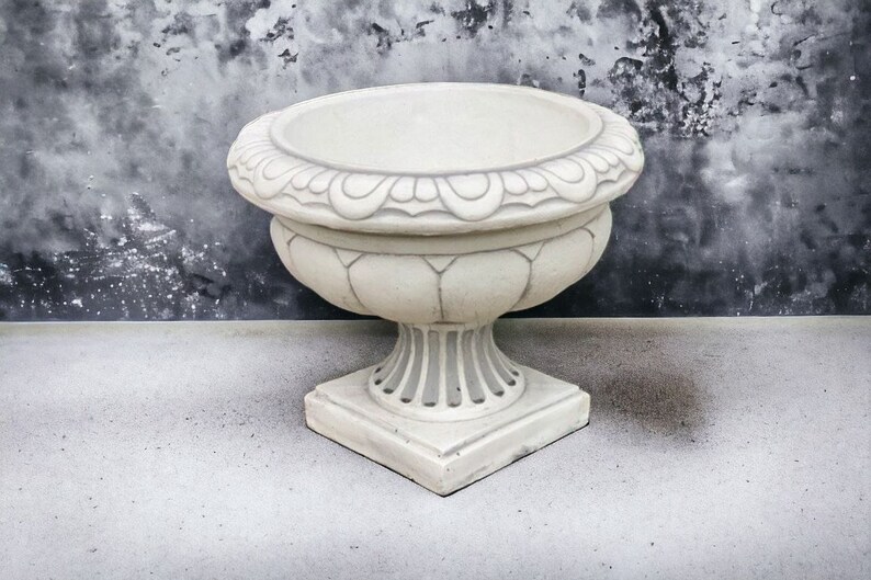 May include: A white ceramic planter with a decorative rim and a square base. The planter is sitting on a gray concrete surface.
