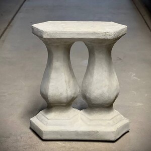May include: A white concrete pedestal with a square top and two rounded columns. The pedestal is made of a rough, textured material.