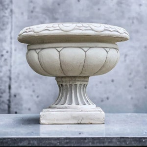 May include: A white ceramic planter with a fluted design and a decorative rim. The planter is sitting on a gray surface.