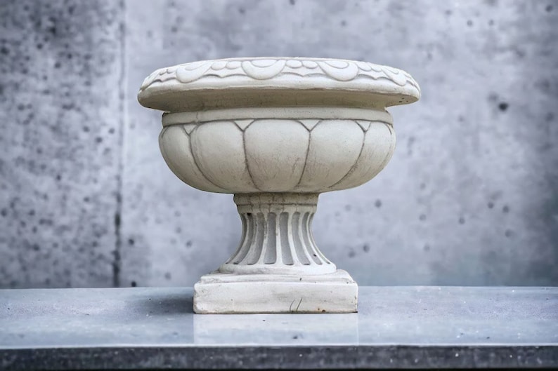 May include: A white ceramic planter with a fluted design and a decorative rim. The planter is sitting on a gray surface.