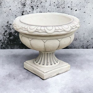 May include: A white ceramic planter with an ornate design. The planter has a wide, round base and a tall, slender neck. It is perfect for displaying flowers or plants.