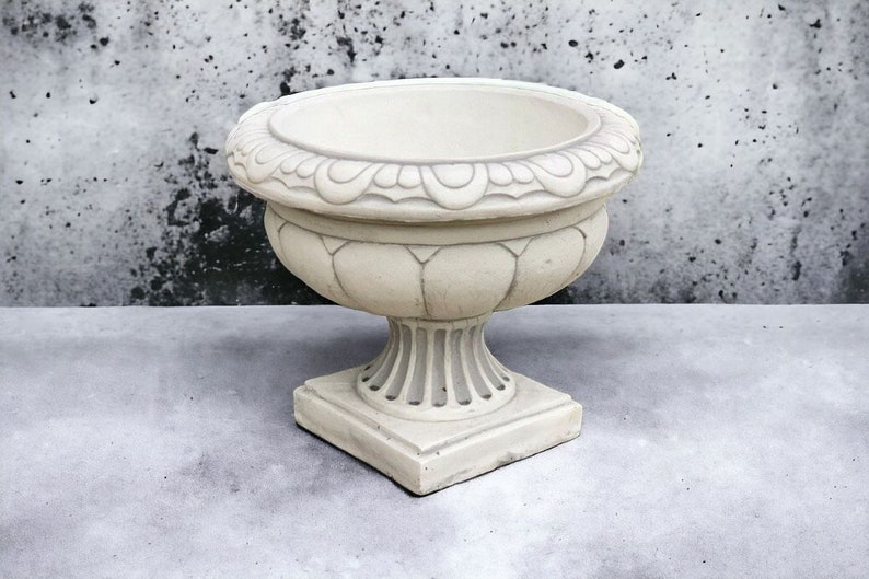 May include: A white ceramic planter with an ornate design. The planter has a wide, round base and a tall, slender neck. It is perfect for displaying flowers or plants.