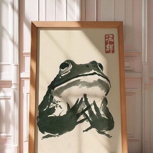 Op de afbeelding: Een ingelijste prent met een inkttekening van een kikker. Het kunstwerk, in een houten lijst, toont een kikker in tinten groen en zwart tegen een lichte achtergrond. De stijl doet denken aan traditionele Japanse kunst.