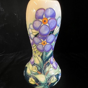 May include: A ceramic vase with a tapered neck and bulbous base, featuring a hand-painted floral pattern of purple flowers with orange centres, green leaves, and white buds against a cream and dark blue background with dark outlines.