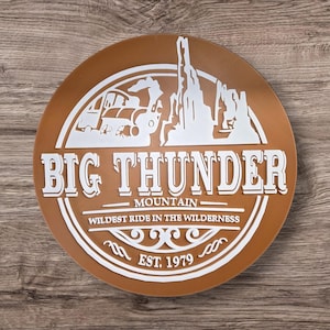 Könnte beinhalten: Ein rundes, braunes Schild mit weißen Buchstaben und Grafiken. Der Text lautet "BIG THUNDER MOUNTAIN" mit einem Bild eines Minenzuges und einer Bergkette. Das Schild enthält auch "WILDEST RIDE IN THE WILDERNESS" und "EST. 1979".