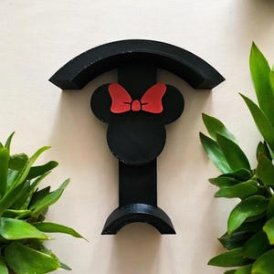 Könnte beinhalten: Schwarzer Wandhaken in Form von Minnie Mouse mit roter Schleife. Der Haken hat eine gebogene Ober- und Unterseite, wobei sich der Minnie Mouse-Kopf in der Mitte befindet. Das Design ist 3D-gedruckt und vor einem neutralen Hintergrund platziert.