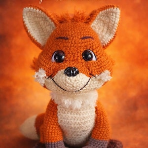 Peut inclure: Une peluche renard au crochet, orange, crème et marron. Le renard a de grands yeux, un nez noir et une bouche souriante. Le texte "Lech'tit Fox par Lachticrochète" est en haut. Le renard est assis.