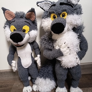 Può includere: Due bambole lupo all'uncinetto fatte a mano con texture in filato grigio e bianco. Entrambe le figure hanno grandi occhi gialli, nasi neri e orecchie a punta. Il lupo più grande ha un petto bianco soffice e una lunga coda folta, mentre il lupo più piccolo ha una macchia bianca liscia sulla pancia. Entrambi sono in piedi con piedi dettagliati simili a zampe.