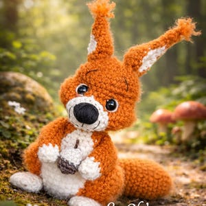 Peut inclure: Un jouet d'écureuil crocheté à la main, orange et blanc, avec un nez noir et de grands yeux, tenant un petit gland. L'image comprend le texte "Lachticrochète" et "Le Ch'tit roux". L'écureuil est placé sur un fond de forêt flou.