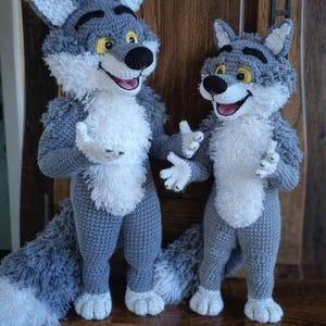 Op de afbeelding: Twee handgemaakte gehaakte wolfpoppen. De poppen zijn grijs met witte vacht op hun borst en gezicht. Ze hebben zwarte neuzen, gele ogen en roze monden. De poppen staan rechtop met hun armen uitgestrekt en hebben lange, pluizige staarten.