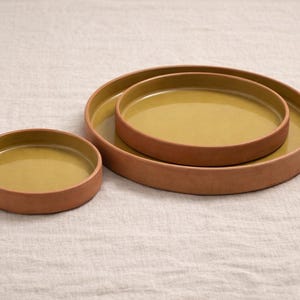 May include: Two round, shallow trays with a matte terracotta exterior and a glossy, mustard-yellow interior. The larger tray contains a smaller, matching tray nested inside, while a third, smaller tray sits separately to the left.