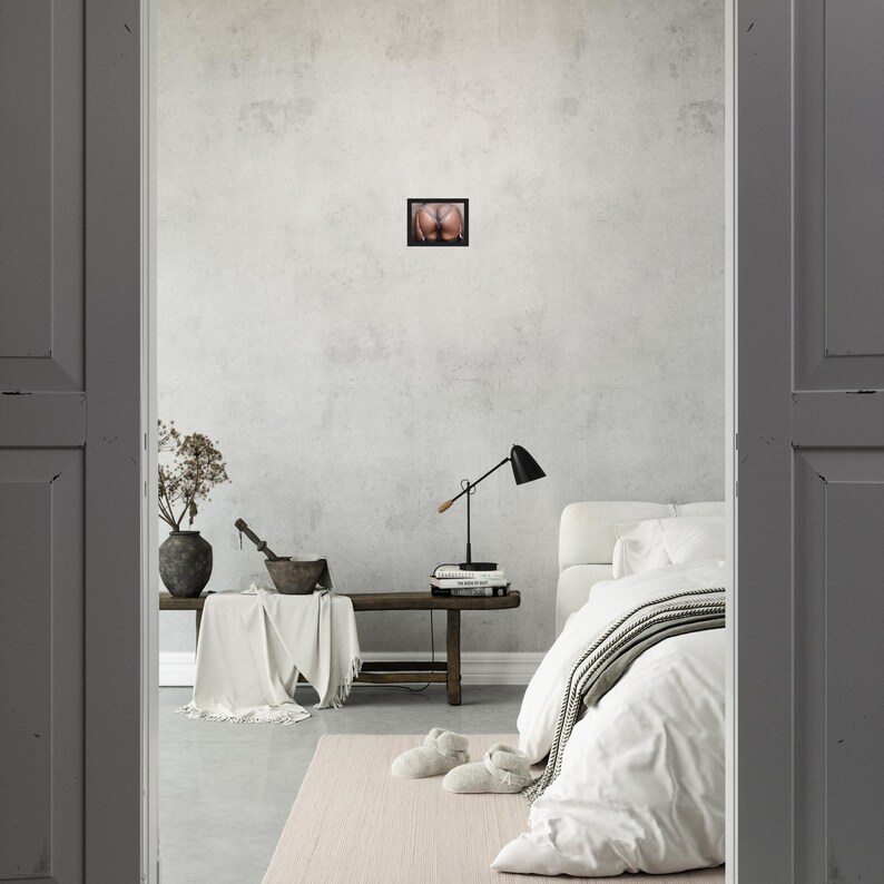 Op de afbeelding: Een ingelijste foto van de billen van een persoon is gecentreerd op een lichtgrijze muur. De foto staat in een zwarte lijst. De kamer heeft een bed met wit beddengoed, een houten bank en een zwarte bureaulamp.