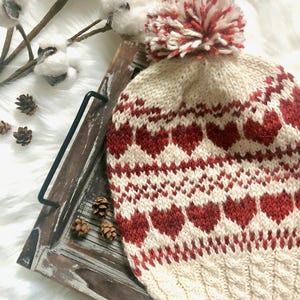 Puede incluir: Un gorro de punto blanco con un patrón de corazones rojos y un pompón blanco y rojo en la parte superior. El gorro está sobre una bandeja de madera con piñas.
