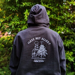 May include: A black hooded sweatshirt with a white graphic of a chef holding a large plant and the text "HASE MIT ALLES?" on the back.
