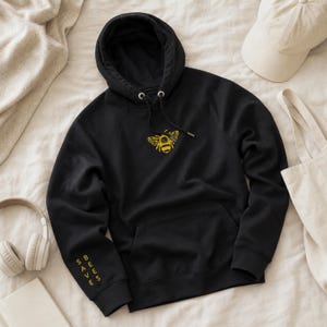 May include: A black hooded sweatshirt featuring a yellow embroidered bee graphic on the center chest and the text SAVE BEES printed in yellow vertically along the right sleeve. The hoodie includes a drawstring hood and a front kangaroo pocket.