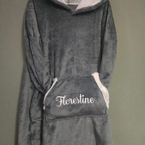Peut inclure: Une couverture à capuche grise surdimensionnée avec une capuche et une poche doublées de sherpa blanc. La poche porte le nom "Florestine" brodé en écriture blanche. La couverture est en matière douce et moelleuse.