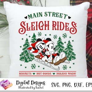 Puede incluir: Una almohada blanca con un diseño rojo y verde que presenta a Mickey y Minnie Mouse montando un trineo en una escena de invierno. El texto "Main Street Sleigh Rides" está en la parte superior de la almohada. El texto "Beignets Hot Cocoa Holiday Magic" está en la parte inferior de la almohada.