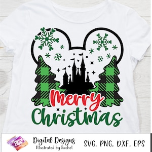 Könnte beinhalten: Ein weißes T-Shirt mit einem schwarz-grünen Karomuster, das eine Silhouette eines Schlosses, zwei Bäume und Schneeflocken zeigt. Das Design hat die Form von Mickey-Maus-Ohren. Der Text "Merry Christmas" ist in roten und grünen Buchstaben unter dem Design geschrieben.