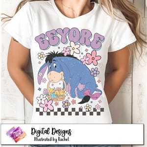 以下が含まれることがあります： ウッディ・ウッドペッカーのキャラクター、イーヨーが描かれた白いTシャツ。イーヨーは、ヒヨコの入ったバスケットを持っています。グラフィックは、ピンクとパープルの花と白黒の市松模様で囲まれています。テキスト「EEYORE」は、パープルとピンクのレトロなフォントで書かれています。