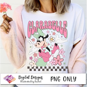 May include: A white t-shirt with a pink and green cartoon character wearing a pink and green outfit. The character is surrounded by pink flowers and the text "ClaraBelle" is written in pink letters.