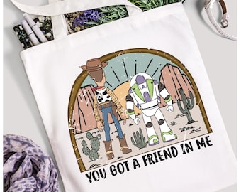 May include: A white tote bag with a vintage-style graphic of Woody and Buzz Lightyear from Toy Story. The graphic is in a distressed, faded look with a sunset background. The text "You got a friend in me" is printed below the graphic.