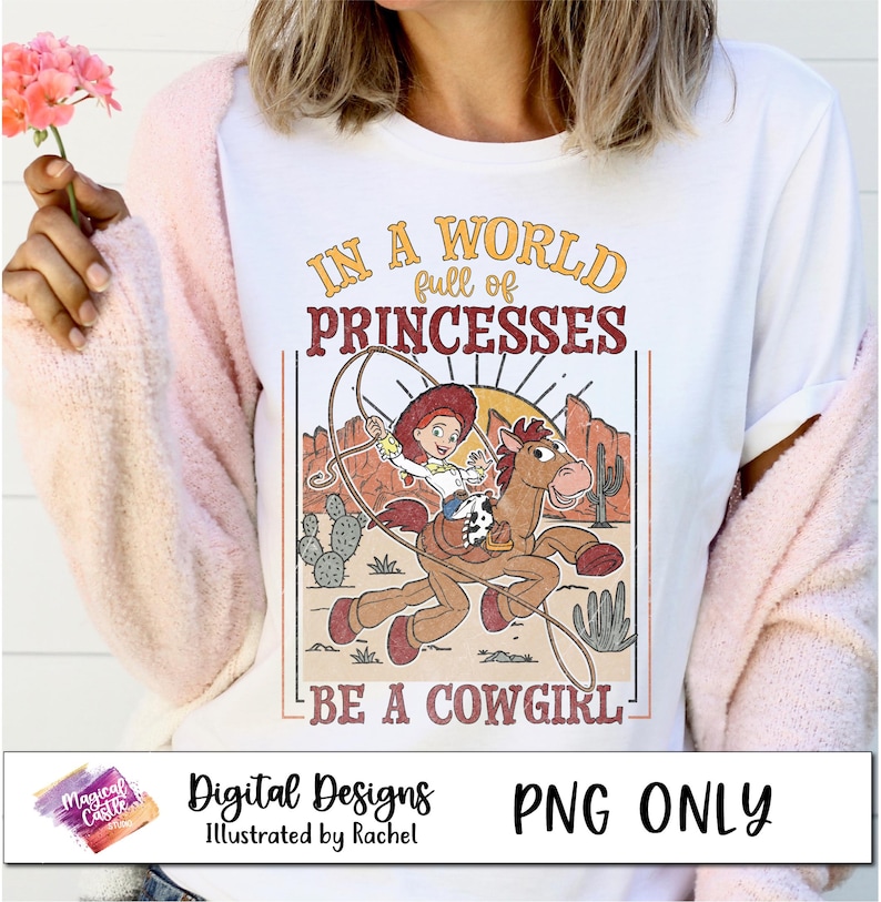 May include: A white t-shirt with a vintage-style graphic featuring a cartoon cowgirl riding a horse. The text reads "In a world full of princesses, be a cowgirl".
